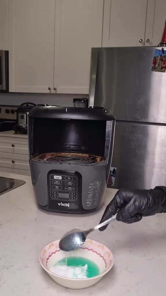 Air fryer jenis buka depan dengan elemen pemanas berkerak minyak keperangan, tangan bersarung hitam memegang sudu dan mangkuk pes pencuci