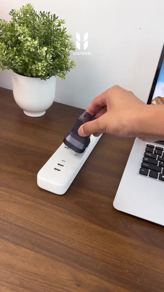 Tangan sedang mencucuk pengecas nipis UGREEN Nexode Air ke extension socket putih di atas meja kayu sebelah laptop