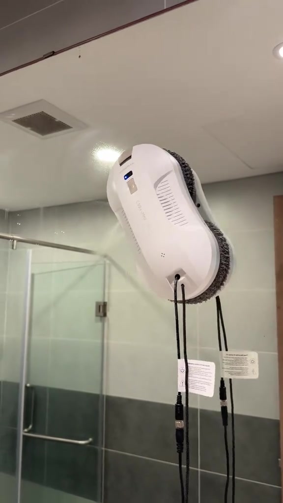 Robot lau kính màu trắng bám trên vách kính phòng tắm, dây điện buông xuống