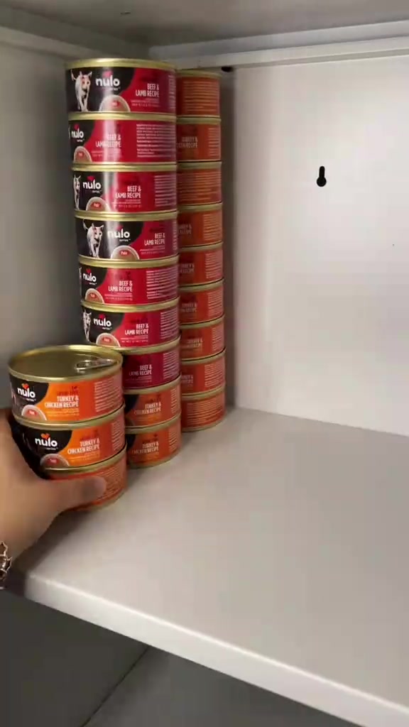 Tin makanan kucing tersusun di rak terbuka, dengan tangan sedang meletakkan satu tin.