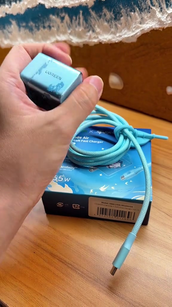Pengecas 65W di tangan dengan kabel USB-C dan kotak produk di belakang