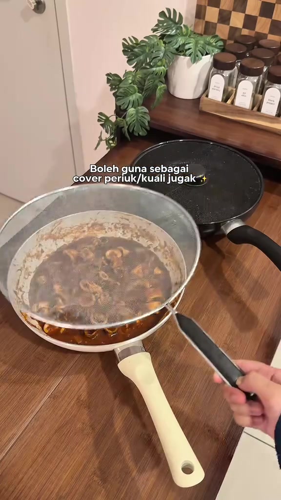 Dua penutup jaring berlainan saiz di atas kuali, satu sedang diangkat dengan tangan.