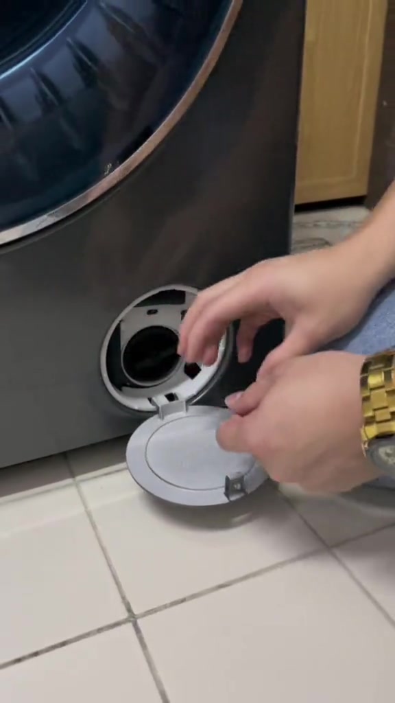 Tangan membuka panel servis kecil di bahagian bawah hadapan mesin basuh untuk akses ke penapis pam.