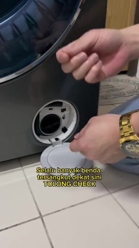Tangan menunjuk bukaan penapis pam di bahagian bawah mesin basuh; panel servis terbuka.